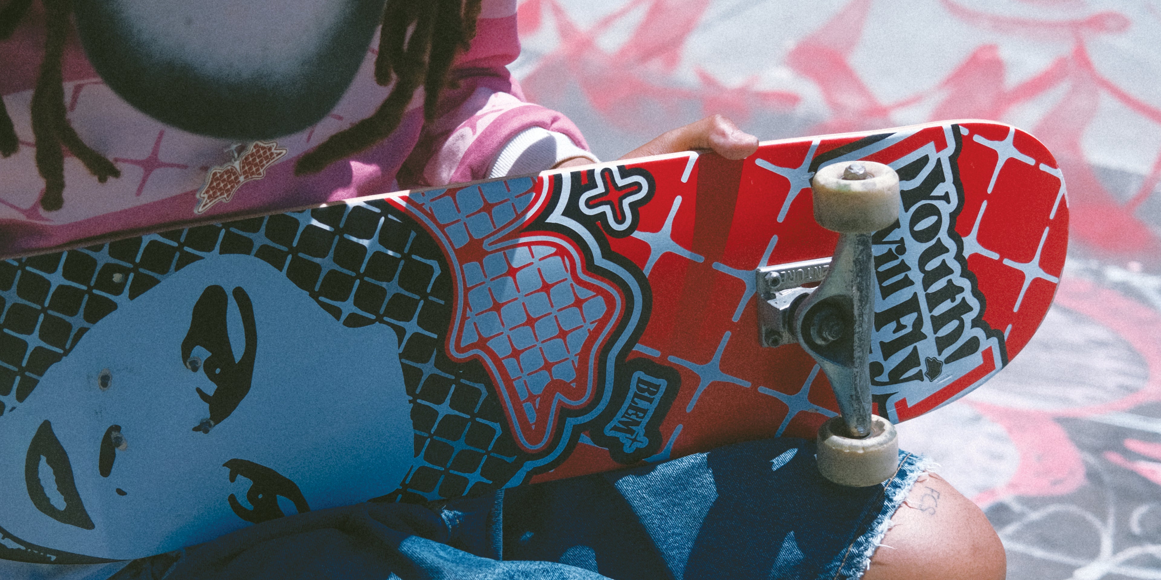 Close-up of a skateboard featuring bold red and black graphic designs with the Blem logo and the text 'Youth Will Fly.' A person wearing a pink sweatshirt and ripped jeans holds the skateboard, highlighting Blem’s creative blend of streetwear culture and skateboarding aesthetics