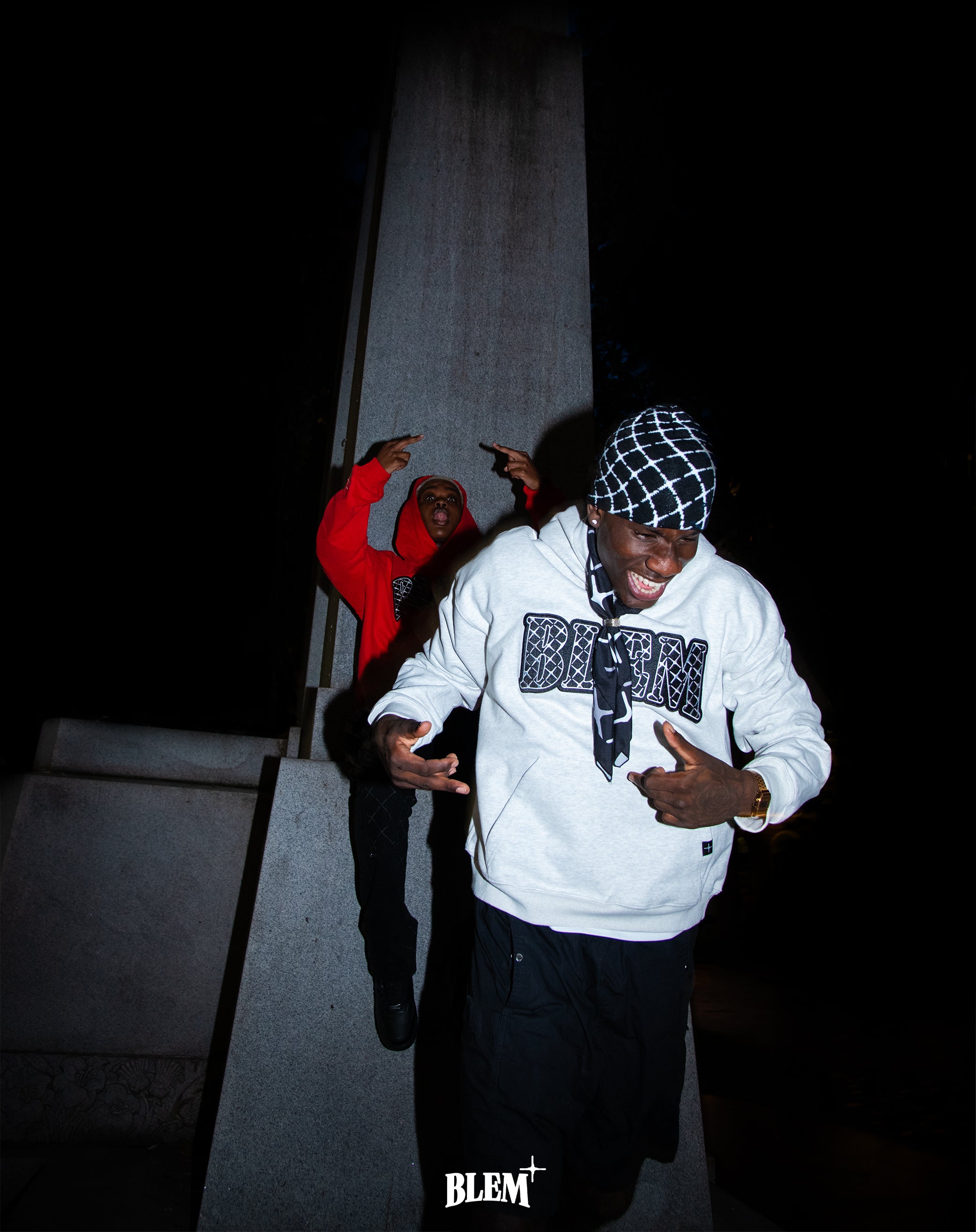 Two stylish young men showcase bold streetwear by Blem, featuring a gray hoodie with the Blem logo and a red hoodie with intricate designs. The scene is set against a striking urban backdrop at night, emphasizing Montreal's vibrant street culture and edgy fashion vibes.